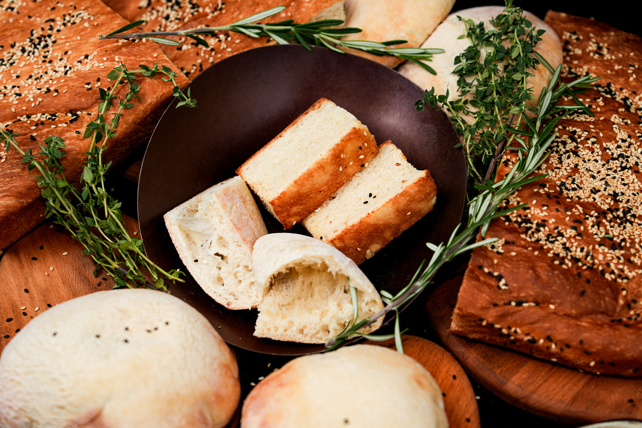 A basket of sliced bread surrounded by seeded flatbreads and herbs