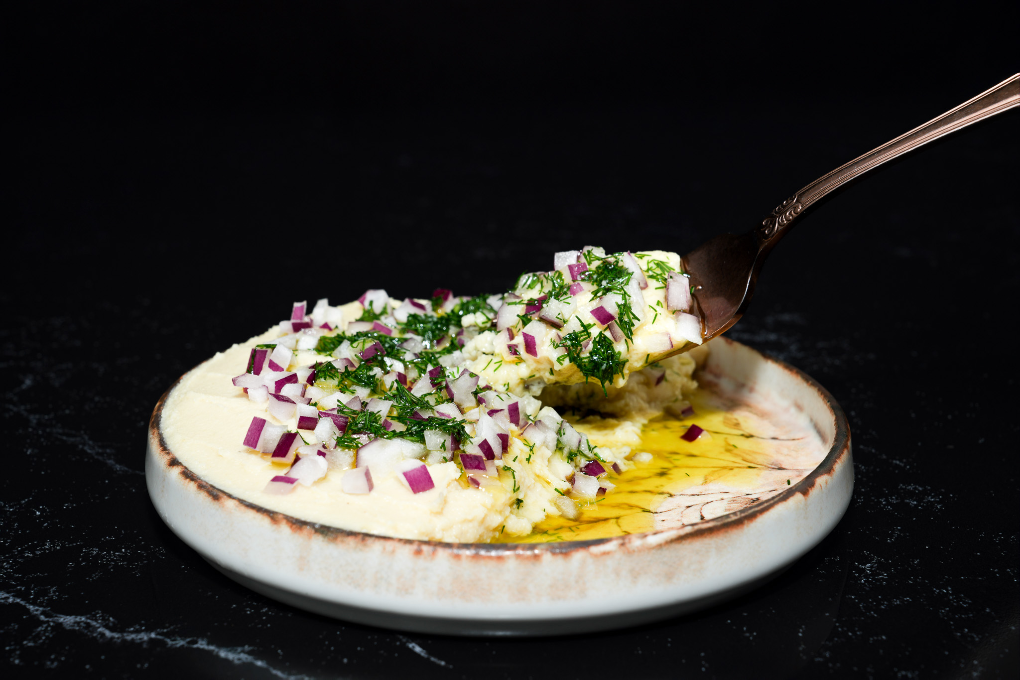A spoon lifting creamy dip topped with chopped herbs and red onion