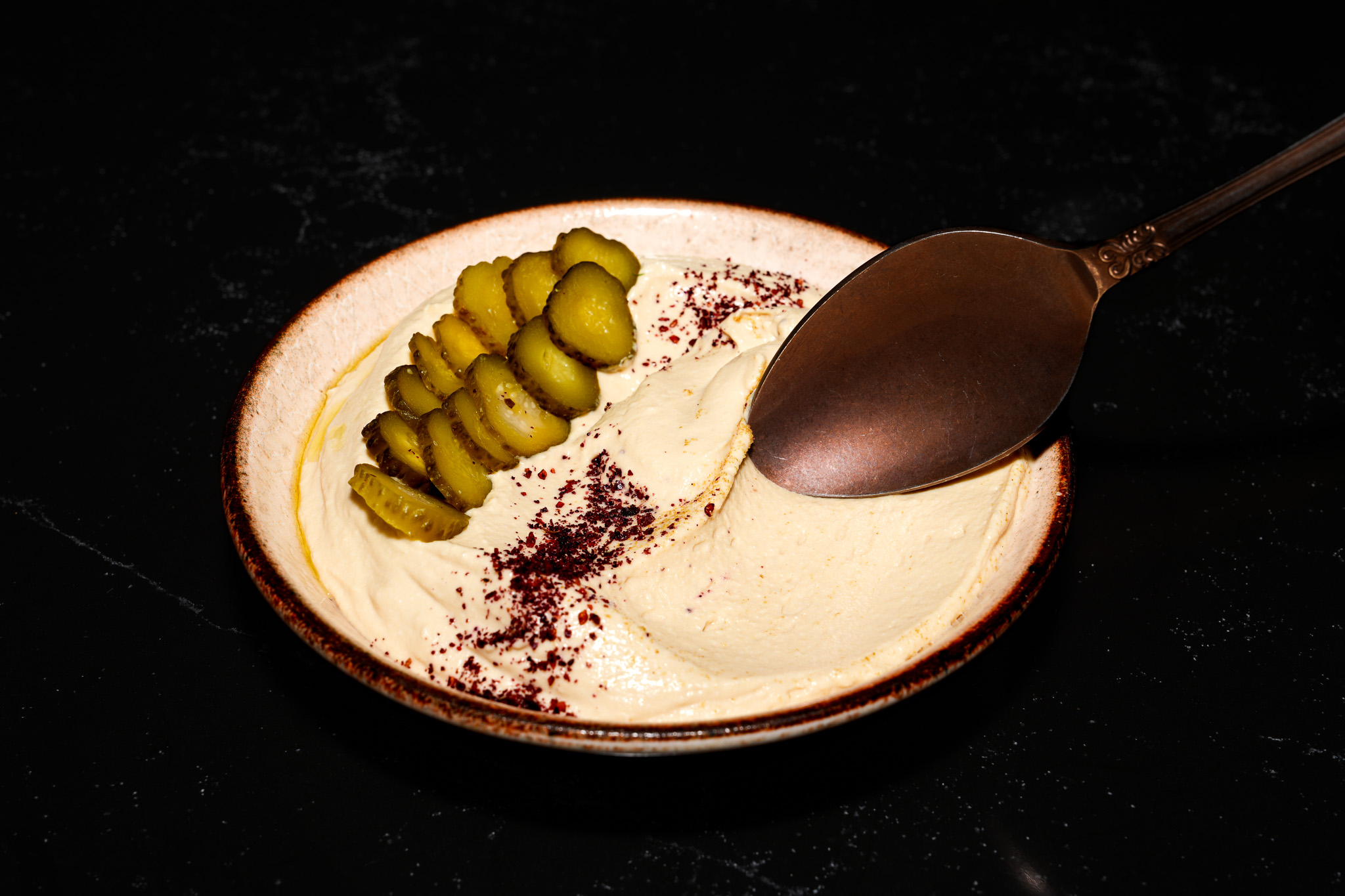 Smooth hummus served with sliced pickles and a spoon