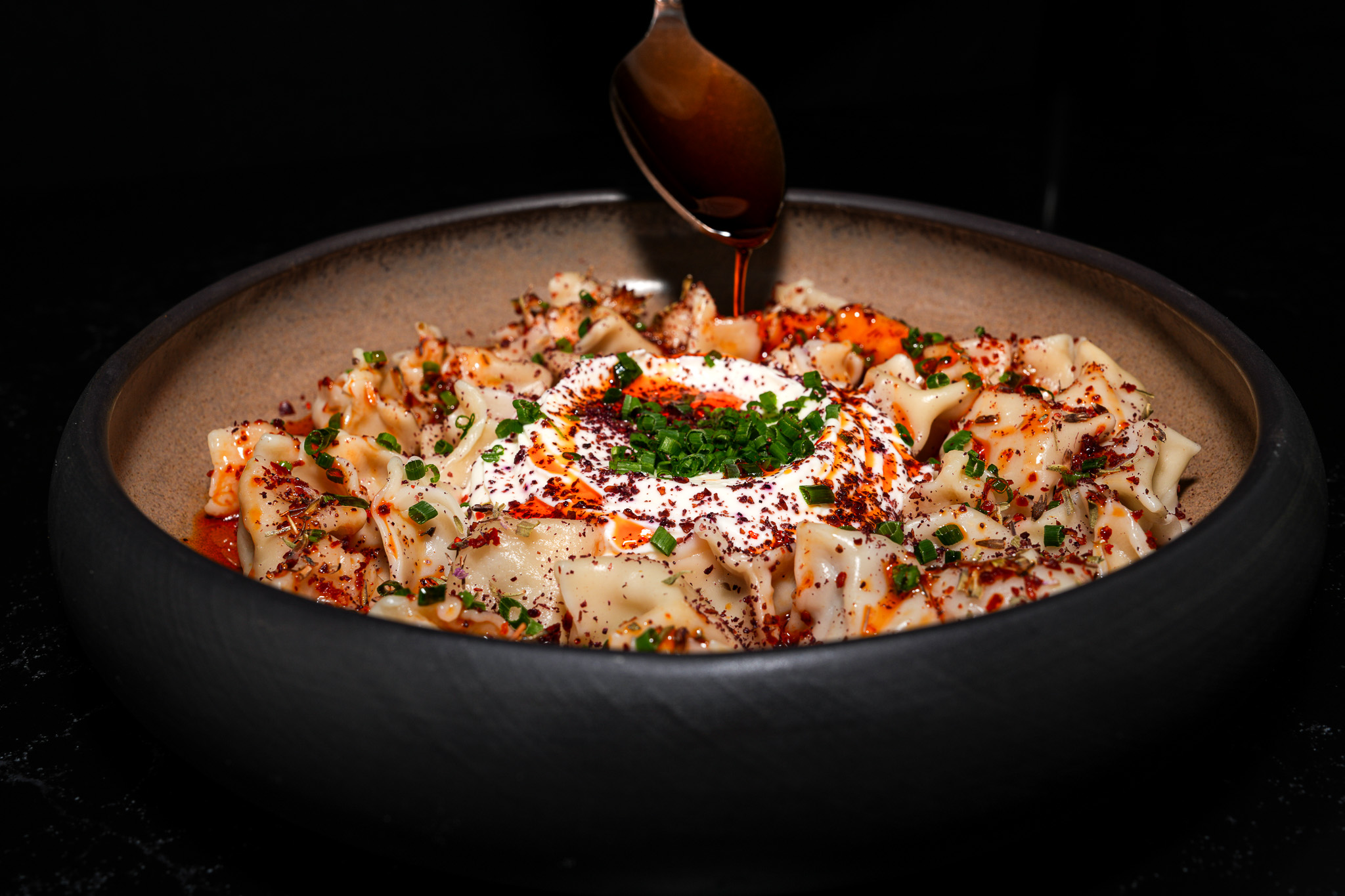 Sauce being spooned over manti with yogurt and herbs