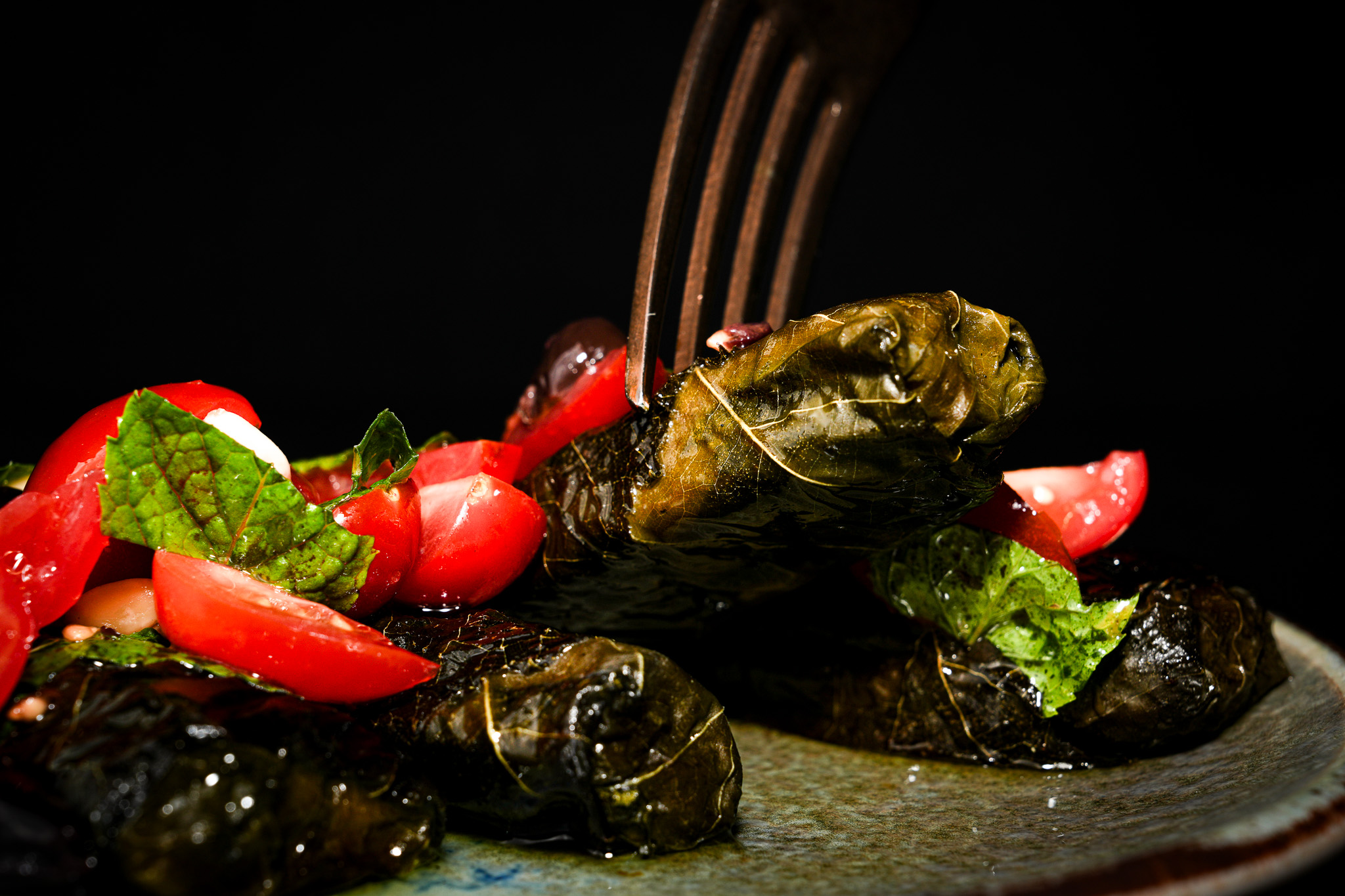 A fork lifting a stuffed vine leaf beside tomatoes and herbs